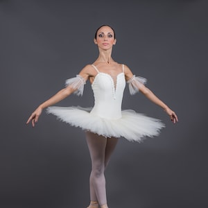 May include: A ballerina in a white tutu and pointe shoes poses on a gray background.