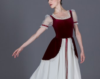 May include: A ballet dancer in a burgundy velvet bodice dress with a white skirt. The dress features lace trim and sheer sleeves. The dancer is wearing ballet shoes and tights, posing against a gray backdrop.