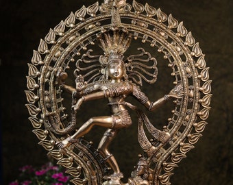 May include: A gold-colored metal sculpture of the Hindu deity Shiva in the Nataraja pose, dancing on a demon. The sculpture is surrounded by a circular halo with intricate details. The sculpture is on a base with the text "Ganeshaartncrafts" on it.