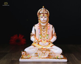Może przedstawiać: Biała marmurowa statua hinduskiego bóstwa Hanuman, siedzącego w medytacyjnej pozie z rękami na kolanach. Ma na sobie żółto-pomarańczową szatę i złotą koronę. Statua stoi na białym marmurowym cokole z złotymi akcentami.