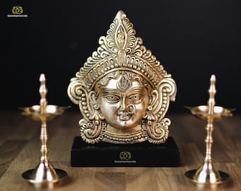 May include: A gold-toned sculpture of a deity's face with ornate headwear, set on a black base. Flanking the sculpture are two brass oil lamps. The logo "Ganeshaartncrafts" is visible.