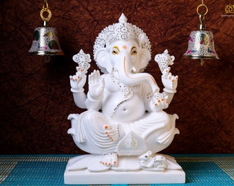 Op de afbeelding: Wit Ganesha-beeld met een gejuwelen kroon en oranje accenten. Het beeld zit op een witte lotusbasis met een kleine witte muis aan zijn voeten. Twee zilveren bellen met kleurrijke ontwerpen hangen boven het beeld.