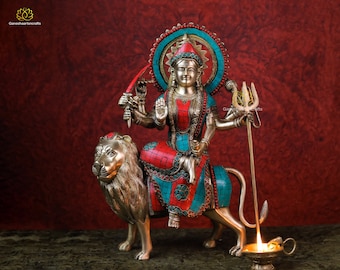 May include: A gold-toned statue of the Hindu goddess Durga riding a lion. The statue is decorated with red and turquoise gemstones. The goddess has four arms and is holding a sword, a trident, and a lotus flower. The statue is set against a red background.