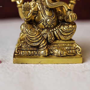 Puede incluir: Una estatua de metal dorado de Ganesha, el dios hind&uacute; de la sabidur&iacute;a y la prosperidad, sentado en una base con intrincados detalles. La estatua est&aacute; en posici&oacute;n sentada con su trompa levantada y sus manos en un gesto de bendici&oacute;n.
