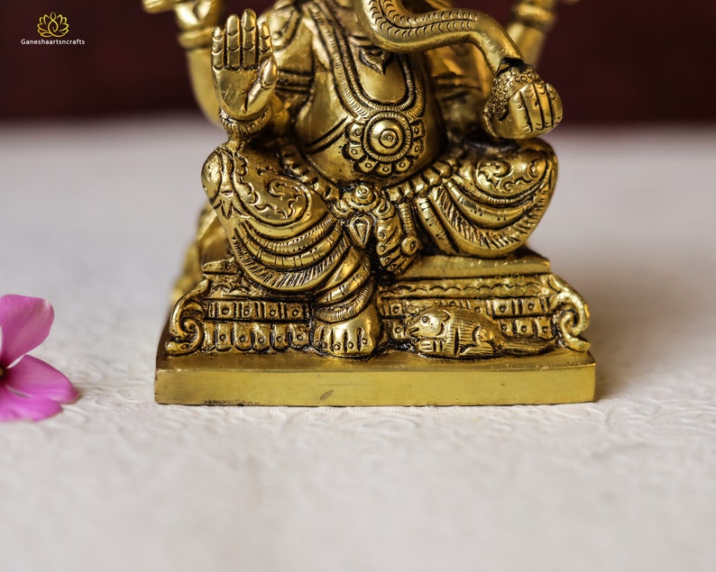 Puede incluir: Una estatua de metal dorado de Ganesha, el dios hind&uacute; de la sabidur&iacute;a y la prosperidad, sentado en una base con intrincados detalles. La estatua est&aacute; en posici&oacute;n sentada con su trompa levantada y sus manos en un gesto de bendici&oacute;n.