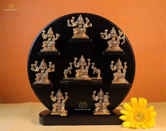 Puede incluir: Un conjunto de ocho figuras de metal dorado de diosas hindúes en un soporte circular negro. Las figuras están dispuestas en círculo y cada una tiene una pose y una expresión diferentes. El soporte tiene un diseño de loto decorativo en la parte inferior.