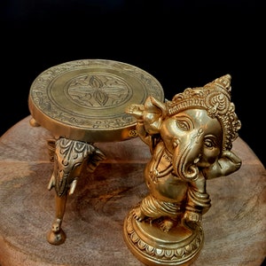 Puede incluir: Un soporte decorativo de metal dorado con una parte superior redonda y tres patas de elefante. El soporte est&aacute; al lado de una estatua de metal dorado de Ganesha, el dios hind&uacute; de los comienzos y eliminador de obst&aacute;culos.