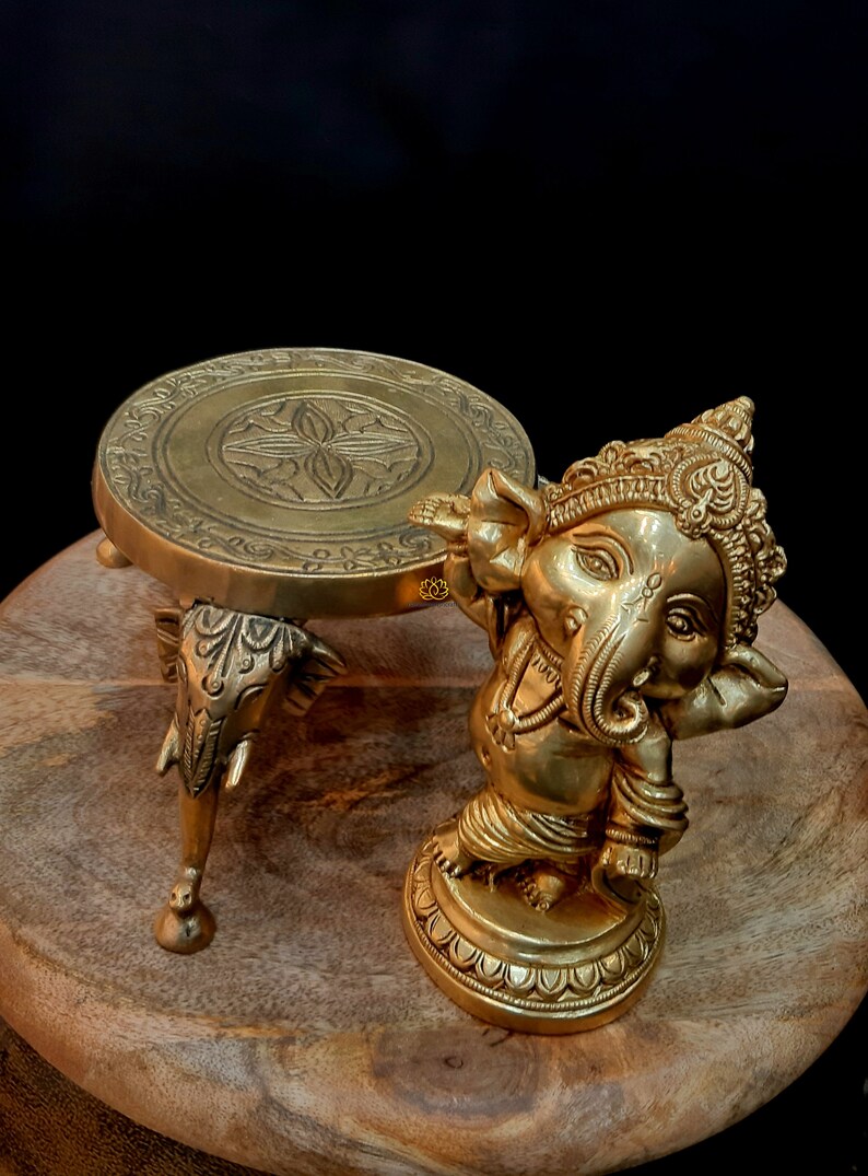 Puede incluir: Un soporte decorativo de metal dorado con una parte superior redonda y tres patas de elefante. El soporte est&aacute; al lado de una estatua de metal dorado de Ganesha, el dios hind&uacute; de los comienzos y eliminador de obst&aacute;culos.