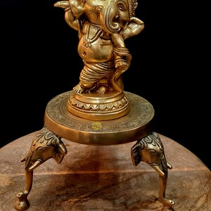 Puede incluir: Una estatua de bronce dorada de Ganesha, el dios hind&uacute; de los comienzos, la prosperidad y la buena fortuna, de pie sobre un soporte decorativo con tres patas de elefante. La estatua est&aacute; sobre una superficie de madera.