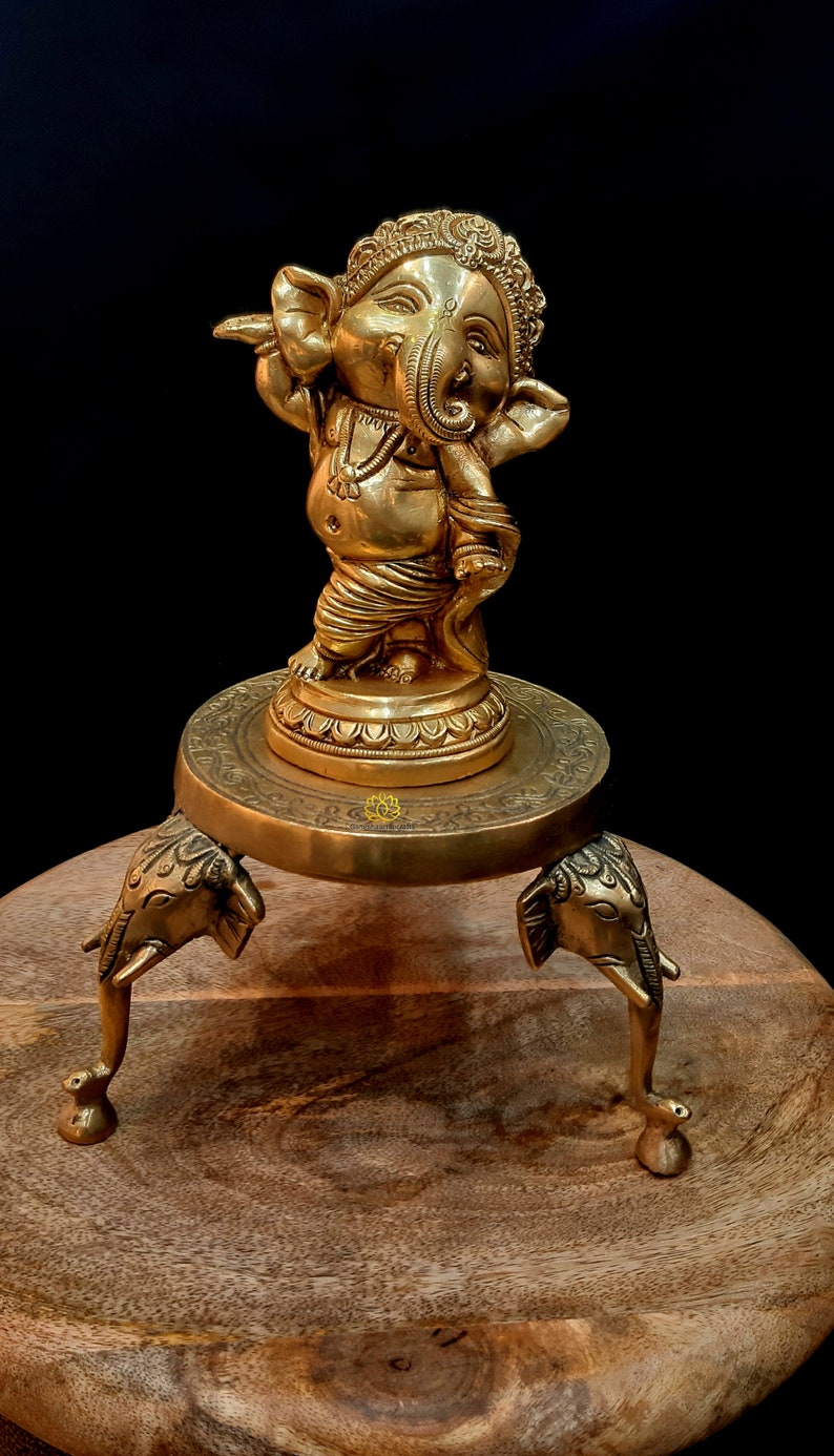 Puede incluir: Una estatua de bronce dorada de Ganesha, el dios hind&uacute; de los comienzos, la prosperidad y la buena fortuna, de pie sobre un soporte decorativo con tres patas de elefante. La estatua est&aacute; sobre una superficie de madera.