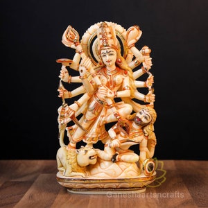 May include: A white and gold statue of a Hindu goddess with multiple arms, standing on a lion. The goddess is holding various weapons and symbols. The statue is on a white base with gold accents.