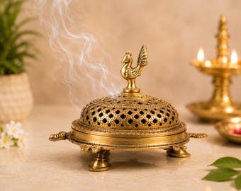 May include: A gold-colored metal incense burner shaped like a turtle with four legs and a tail. The dome-shaped lid features an intricate lattice pattern and a central peacock figurine on top, with thin wisps of white smoke rising from the vents.
