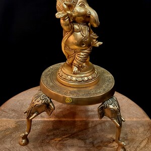 Puede incluir: Una estatua de bronce dorado de Ganesha, el dios hind&uacute; de la sabidur&iacute;a y la prosperidad, de pie sobre una base decorativa con tres patas de elefante. La estatua est&aacute; intrincadamente detallada y presenta un dise&ntilde;o tradicional.