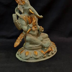 Op de afbeelding: Een klein, groen en gouden Ganesha-beeldje dat op een lotusbasis zit. Het beeldje is gemaakt van metaal en heeft ingewikkelde details.