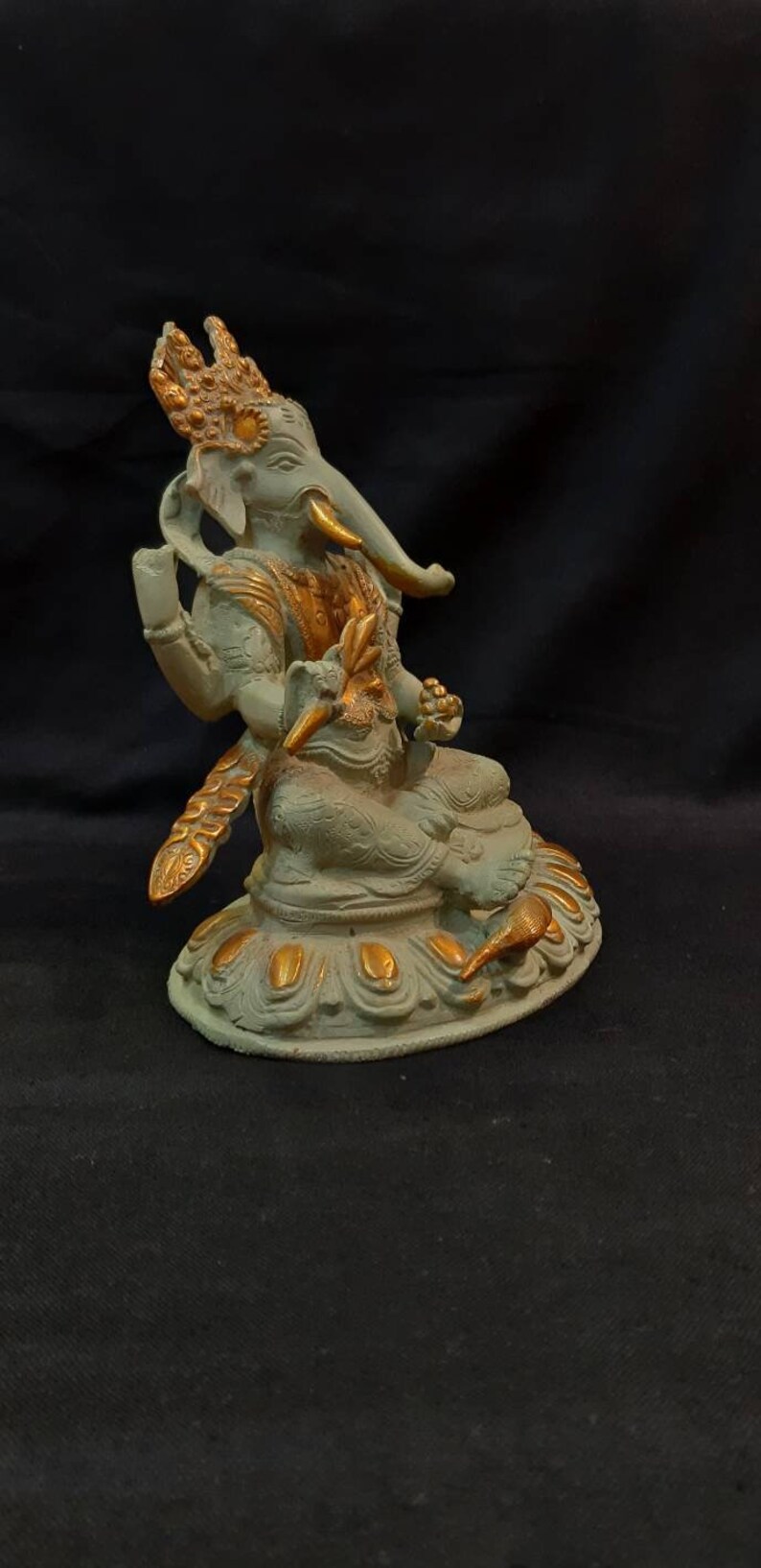 Op de afbeelding: Een klein, groen en gouden Ganesha-beeldje dat op een lotusbasis zit. Het beeldje is gemaakt van metaal en heeft ingewikkelde details.