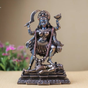 May include: A detailed statue of the Hindu goddess Kali, depicted with multiple arms, holding a curved sword and a bowl. The statue is dark blue with bronze accents, standing on a figure, and adorned with a garland of skulls. The base is rectangular.