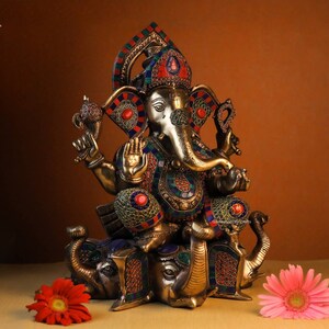 May include: A gold-colored statue of Ganesha, the Hindu god of wisdom and prosperity, sitting on a base of three elephants. The statue is decorated with colorful gemstones and intricate details.