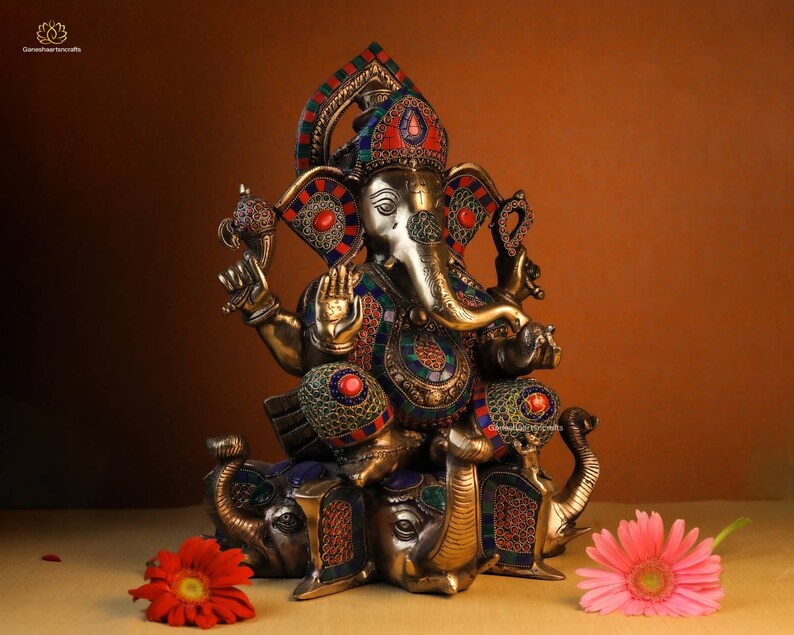 May include: A gold-colored statue of Ganesha, the Hindu god of wisdom and prosperity, sitting on a base of three elephants. The statue is decorated with colorful gemstones and intricate details.