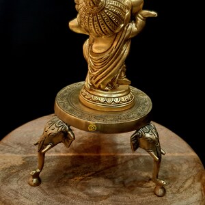 Puede incluir: Una estatua dorada de una deidad, posiblemente Krishna, tocando la flauta, de pie sobre una base redonda con tres patas de elefante. La estatua est&aacute; sobre una superficie de madera.