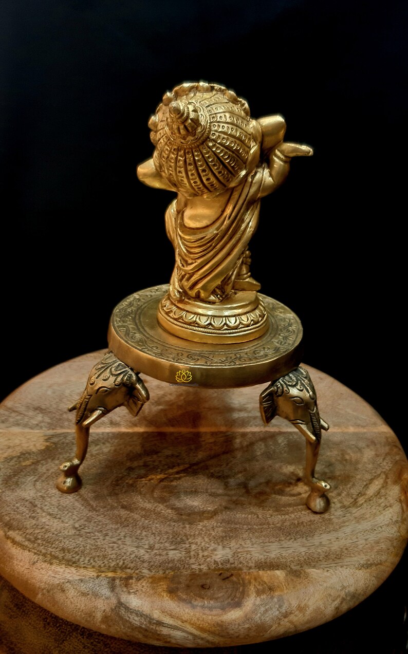 Puede incluir: Una estatua dorada de una deidad, posiblemente Krishna, tocando la flauta, de pie sobre una base redonda con tres patas de elefante. La estatua est&aacute; sobre una superficie de madera.