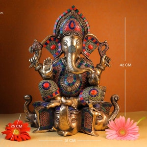 May include: A gold-toned statue of Ganesha, the Hindu god of wisdom and prosperity, sitting on two elephants. The statue is decorated with colorful gemstones and intricate details. The statue is 42 cm tall, 31 cm wide, and 25 cm deep.