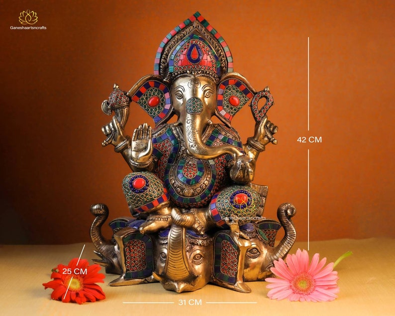 May include: A gold-toned statue of Ganesha, the Hindu god of wisdom and prosperity, sitting on two elephants. The statue is decorated with colorful gemstones and intricate details. The statue is 42 cm tall, 31 cm wide, and 25 cm deep.