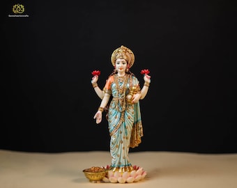 May include: A white and gold statue of Lakshmi, the Hindu goddess of wealth and prosperity. She is standing on a lotus flower base and holding a lotus flower in each hand. She is wearing a blue and white sari with gold accents.