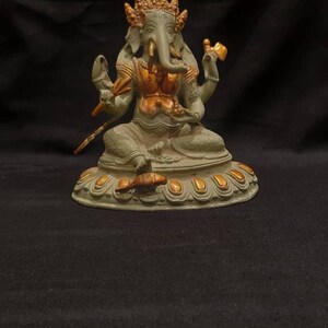 Op de afbeelding: Een groen en gouden Ganesha-beeld met vier armen, zittend op een lotusbasis. Het beeld is gemaakt van metaal en heeft een gedetailleerd ontwerp.