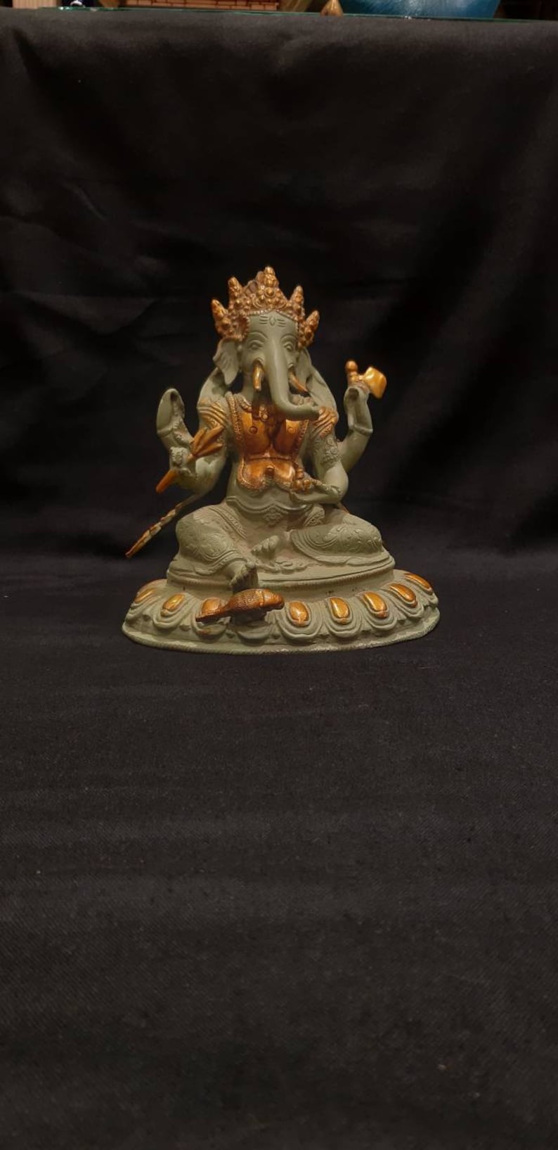 Op de afbeelding: Een groen en gouden Ganesha-beeld met vier armen, zittend op een lotusbasis. Het beeld is gemaakt van metaal en heeft een gedetailleerd ontwerp.