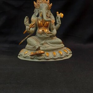 Op de afbeelding: Een groen en gouden Ganesha-beeld met een kroon en vier armen. Het beeld zit op een ronde basis met gouden accenten.