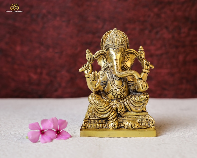 Puede incluir: Una estatua de bronce dorada de la deidad hind&uacute; Ganesha, sentado con la trompa levantada y sosteniendo un objeto peque&ntilde;o en su mano. La estatua est&aacute; sobre una base cuadrada y tiene detalles intrincados.