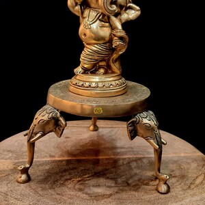 Puede incluir: Una estatua de bronce dorada de Ganesha, el dios hind&uacute; de la sabidur&iacute;a y la prosperidad, de pie sobre un soporte decorativo con tres patas de elefante. El soporte es de madera y tiene una parte superior redonda.
