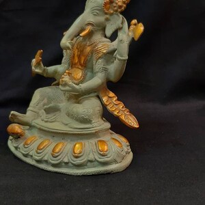Op de afbeelding: Een groen en gouden Ganesha-beeld met een kroon en een decoratieve basis. Het beeld staat op een zwarte ondergrond.