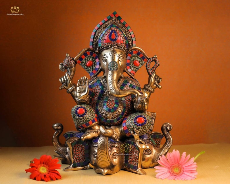 May include: A gold-colored statue of Ganesha, the Hindu god of wisdom and prosperity, sitting on a double elephant base. The statue is decorated with colorful gemstones and intricate details.