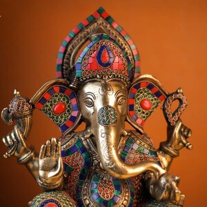 May include: A gold-colored Ganesha statue with intricate details and colorful gemstone accents. The statue is adorned with red, blue, and green gemstones, and the elephant's trunk is raised in a gesture of blessing.