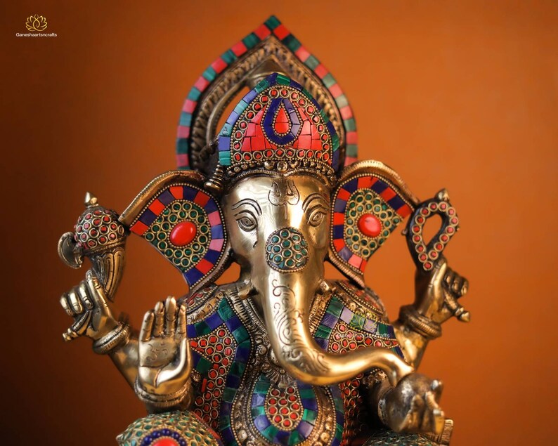 May include: A gold-colored Ganesha statue with intricate details and colorful gemstone accents. The statue is adorned with red, blue, and green gemstones, and the elephant's trunk is raised in a gesture of blessing.