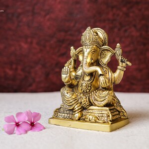 Puede incluir: Una estatua de metal dorado del dios hind&uacute; Ganesha. La estatua est&aacute; sentada sobre una base y tiene detalles intrincados. La estatua est&aacute; frente a un fondo rojo.