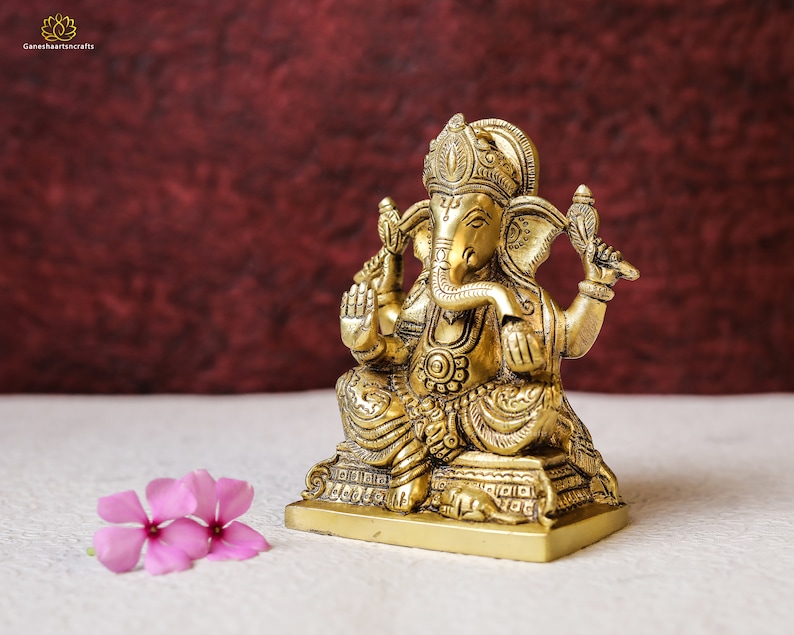Puede incluir: Una estatua de metal dorado del dios hind&uacute; Ganesha. La estatua est&aacute; sentada sobre una base y tiene detalles intrincados. La estatua est&aacute; frente a un fondo rojo.