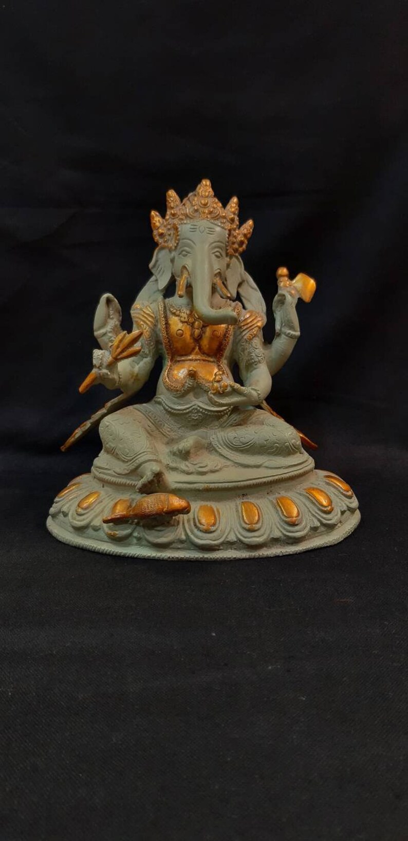Op de afbeelding: Een groen en gouden Ganesha-beeld met een decoratieve basis. Het beeld staat op een zwarte ondergrond.