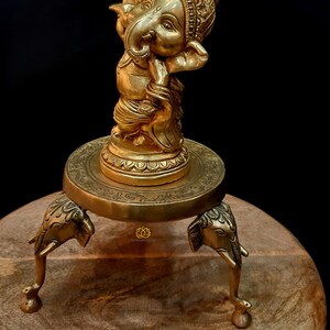 Puede incluir: Una estatua de metal dorado de Ganesha, el dios hind&uacute; de la sabidur&iacute;a y la prosperidad, de pie sobre una base redonda con tres patas de elefante. La estatua est&aacute; sobre una superficie de madera.