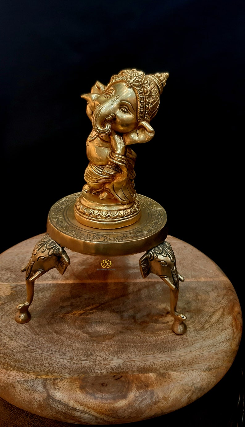 Puede incluir: Una estatua de metal dorado de Ganesha, el dios hind&uacute; de la sabidur&iacute;a y la prosperidad, de pie sobre una base redonda con tres patas de elefante. La estatua est&aacute; sobre una superficie de madera.