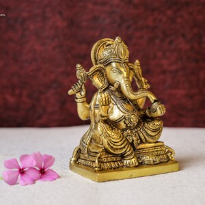 Puede incluir: Una estatua de bronce dorada de Ganesha, el dios hind&uacute; de la sabidur&iacute;a y la prosperidad, sentado en una base con intrincados detalles. La estatua est&aacute; sobre una superficie blanca con flores rosas.