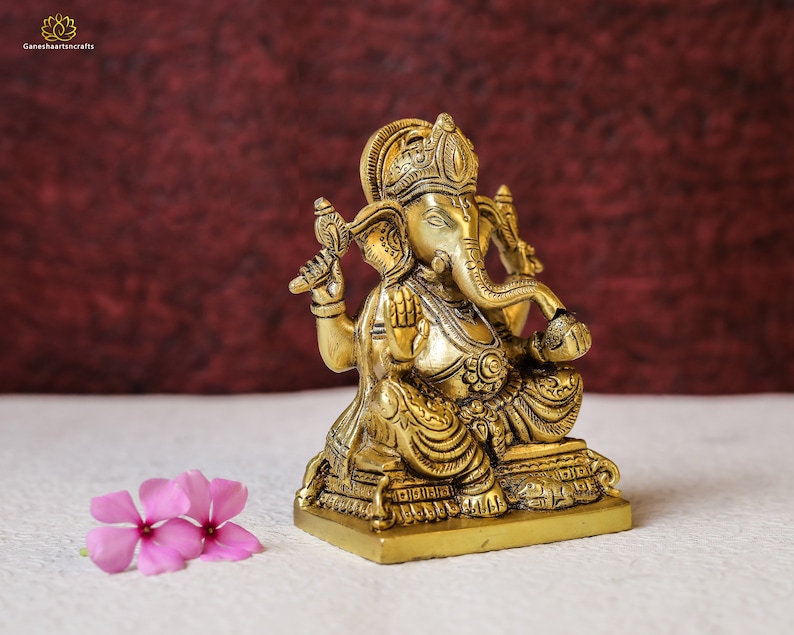 Puede incluir: Una estatua de bronce dorada de Ganesha, el dios hind&uacute; de la sabidur&iacute;a y la prosperidad, sentado en una base con intrincados detalles. La estatua est&aacute; sobre una superficie blanca con flores rosas.