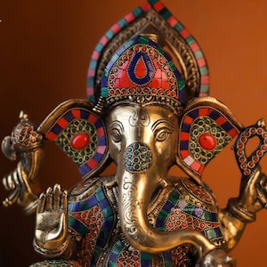 May include: A gold-colored statue of the Hindu deity Ganesha, adorned with intricate details and colorful gemstones. The statue features a large head with an elephant trunk, large ears, and a single tusk. The statue is decorated with red, blue, and green gemstones, and the eyes are inlaid with black stones.