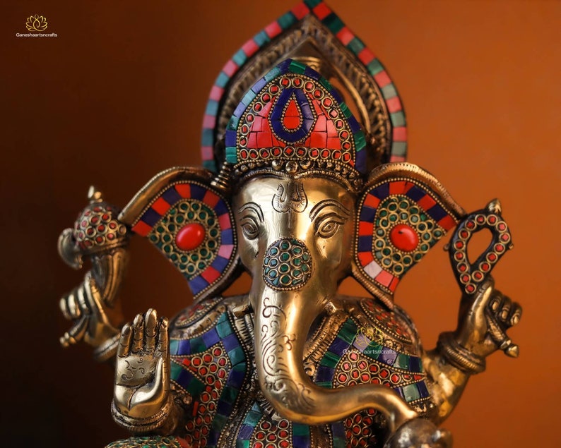 May include: A gold-colored statue of the Hindu deity Ganesha, adorned with intricate details and colorful gemstones. The statue features a large head with an elephant trunk, large ears, and a single tusk. The statue is decorated with red, blue, and green gemstones, and the eyes are inlaid with black stones.