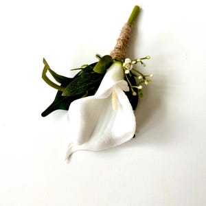 May include: A white calla lily buttonhole with green leaves and small white buds. The stem is wrapped in twine and green tape. The flower is set against a white background.