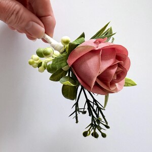 May include: A buttonhole featuring a single dusty rose fabric flower, accented with small green berries, dark green foliage, and thin dark stems. The base of the floral arrangement is wrapped in white floral tape for a clean, finished appearance.