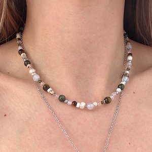 May include: A layered necklace with a silver chain and a small silver charm. The top necklace is made of various colored beads, including brown, green, white, and blue.