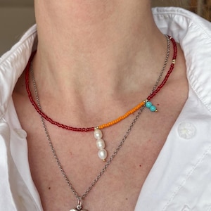 Puede incluir: Un collar de capas con una hebra de cuentas rojas, naranjas y turquesas, una cadena de plata con un colgante de corazón y tres perlas blancas que cuelgan de la hebra de cuentas.