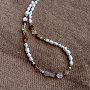 Könnte beinhalten: Eine zarte Halskette mit weißen Perlen und einer Vielzahl farbiger Edelsteine. Die Halskette hat eine goldfarbene Kette und einen Verschluss. Die Edelsteine umfassen Farbtöne von Rosa, Braun, Gelb und Blau. Die Halskette wird auf einer braunen Oberfläche präsentiert.
